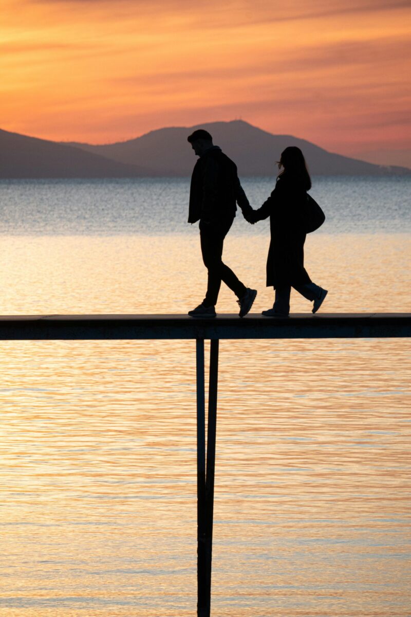 A couple holding hands, silhouetted against a vibrant sunset over a tranquil lake.