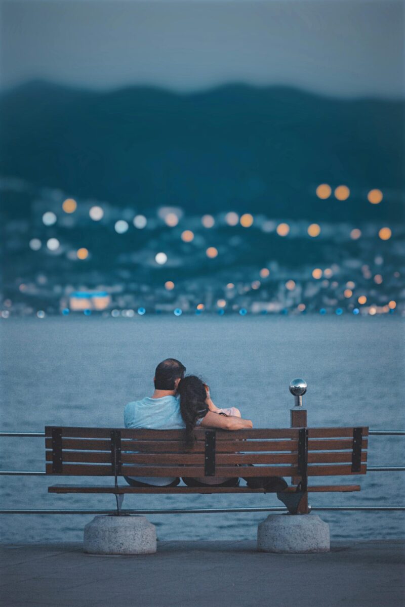 Couple embracing on a bench by a lake with city lights in the background.